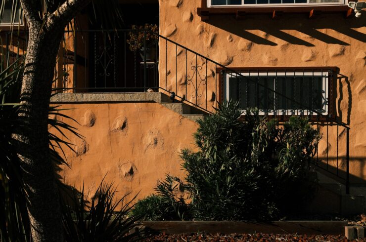 Sunlight and tree shadows fall across an ochre stucco exterior wall and metal stair railing.